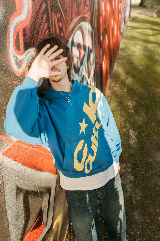 Young man in blue Guilty hoodie leaning against colorful graffiti wall, covering face with hand outdoors.