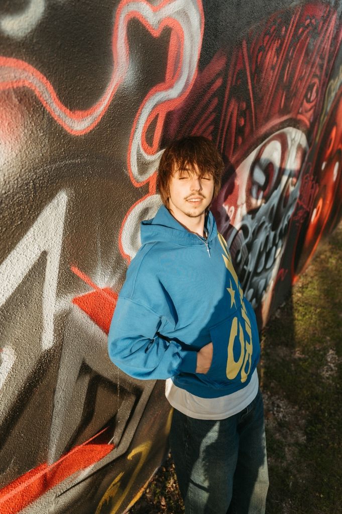 Young man in blue hoodie posing against colorful graffiti wall in urban outdoor setting.