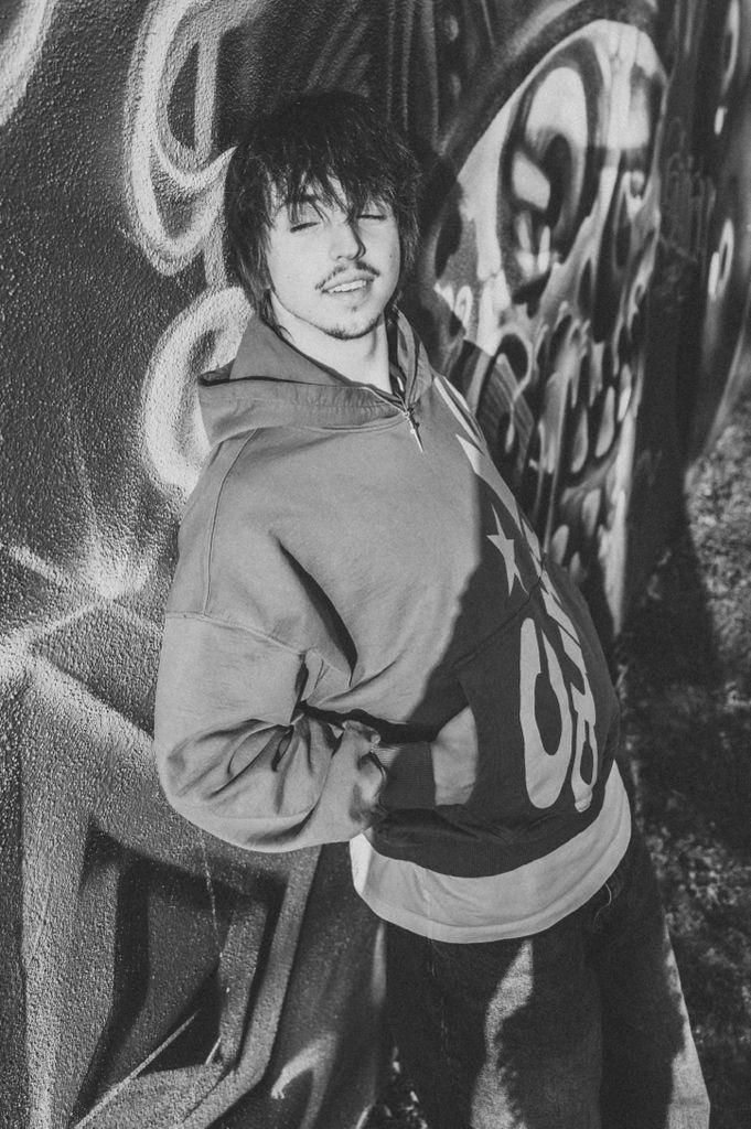 Black and white photo of a young man in a hoodie smiling against a graffiti wall.