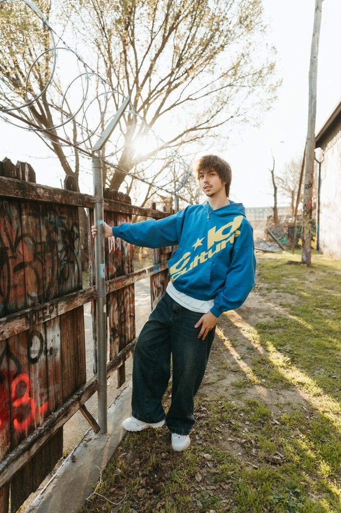 Young man in blue graphic hoodie and baggy jeans leans against graffiti wall outdoors in golden sunlight.