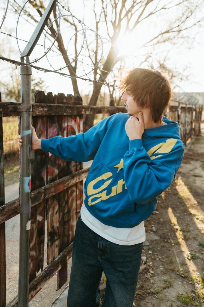 Young person in blue graphic hoodie and jeans leaning on rustic fence with barbed wire, sun flare behind bare trees.