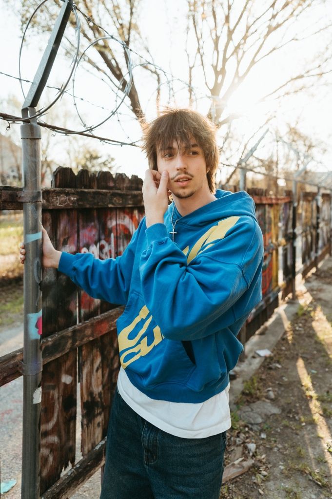 Young man in blue hoodie with yellow design poses by graffiti fence outdoors in sunlight.