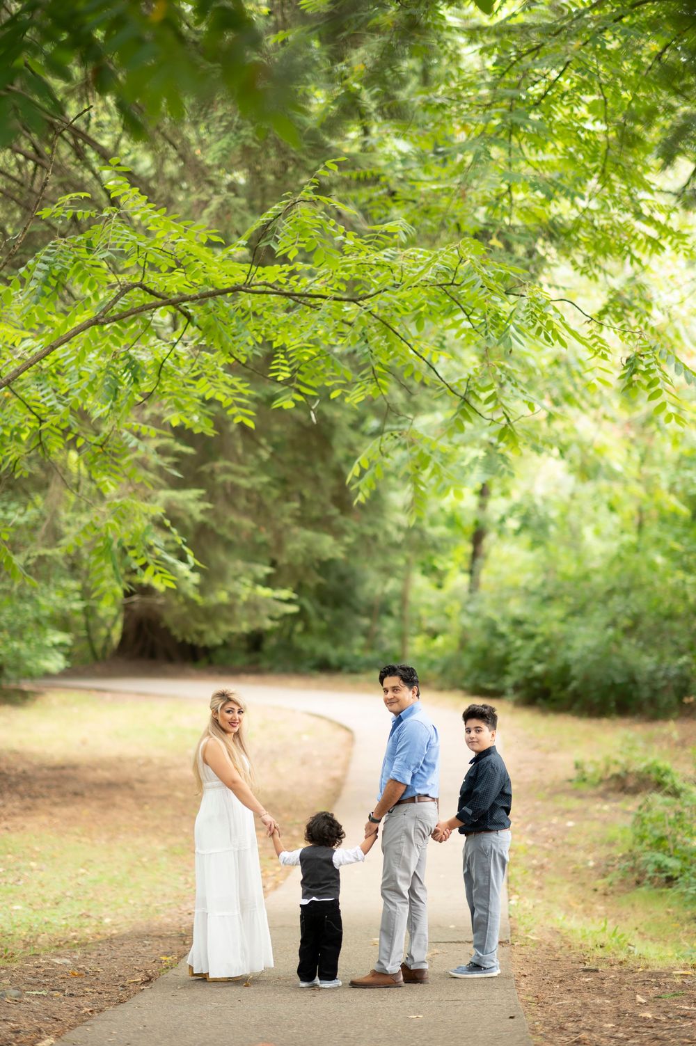 Family of four holding hands on a park path surrounded by lush green trees during an outdoor portrait session.