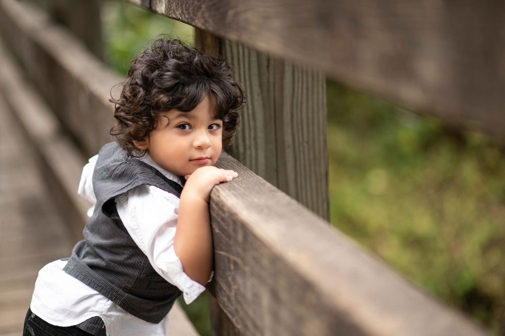 A young curly-haired boy in a vest leans on a wooden fence railing, gazing thoughtfully into the distance.