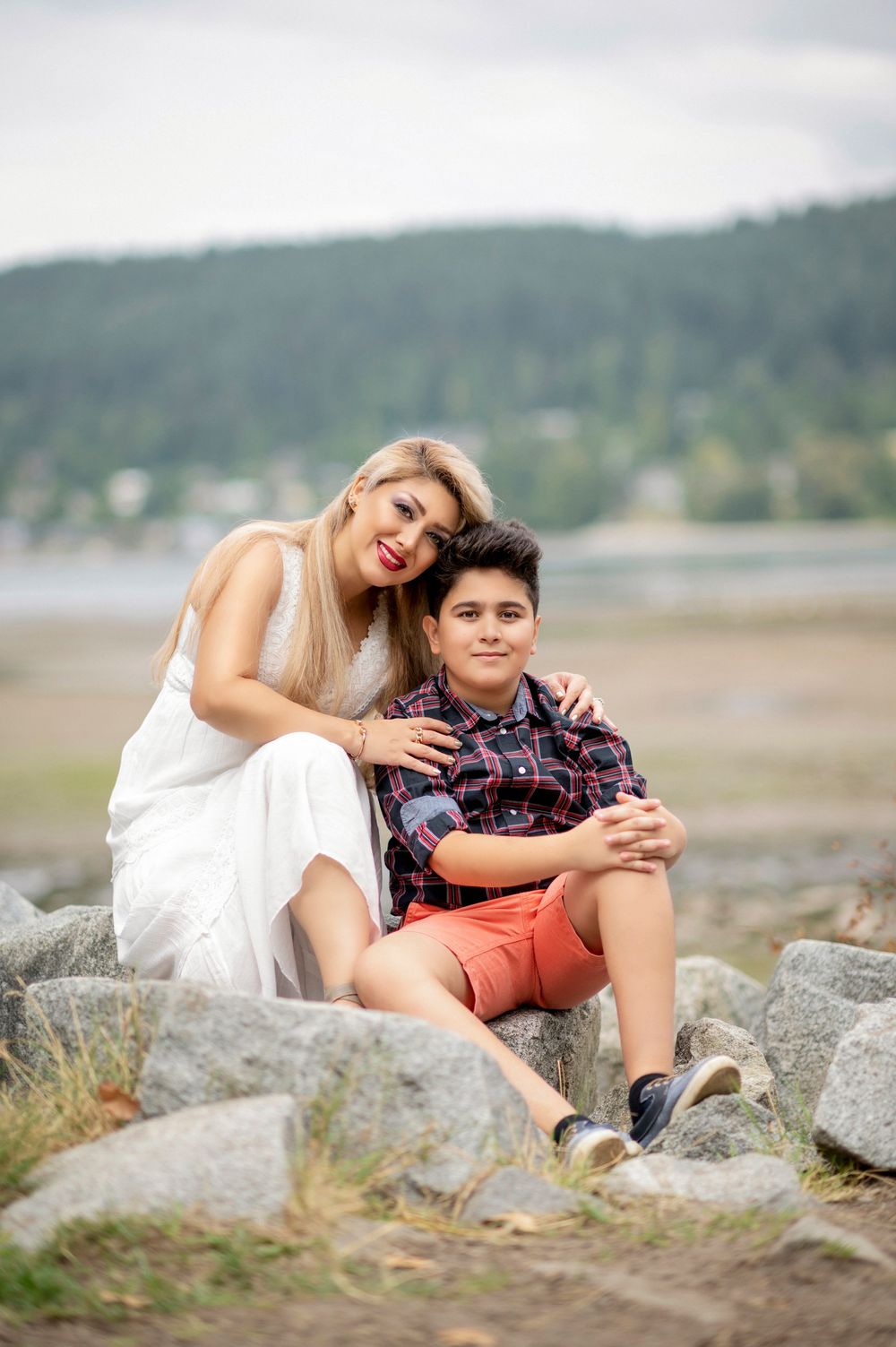 Loving mother in white dress sitting on rocks with her young son at a scenic waterfront location.