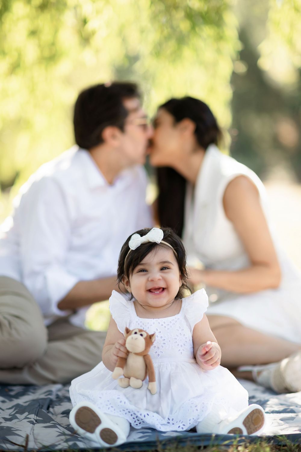 Baby girl in white dress holds teddy bear, smiling, while parents share a kiss outdoors on a blanket.