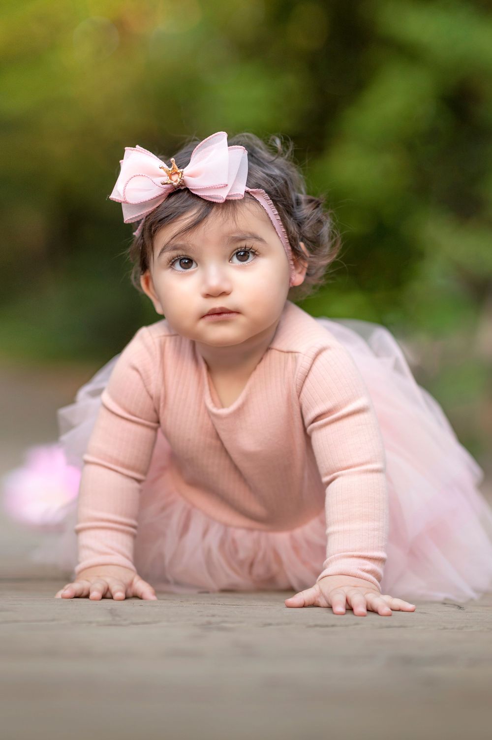 Adorable baby girl in pink tutu dress and bow headband crawling outdoors with green bokeh background.