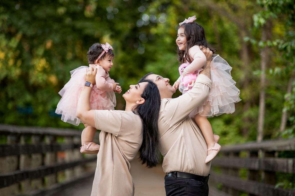 Family portrait with two young girls in pink tutus being lifted joyfully outdoors on a wooden bridge.