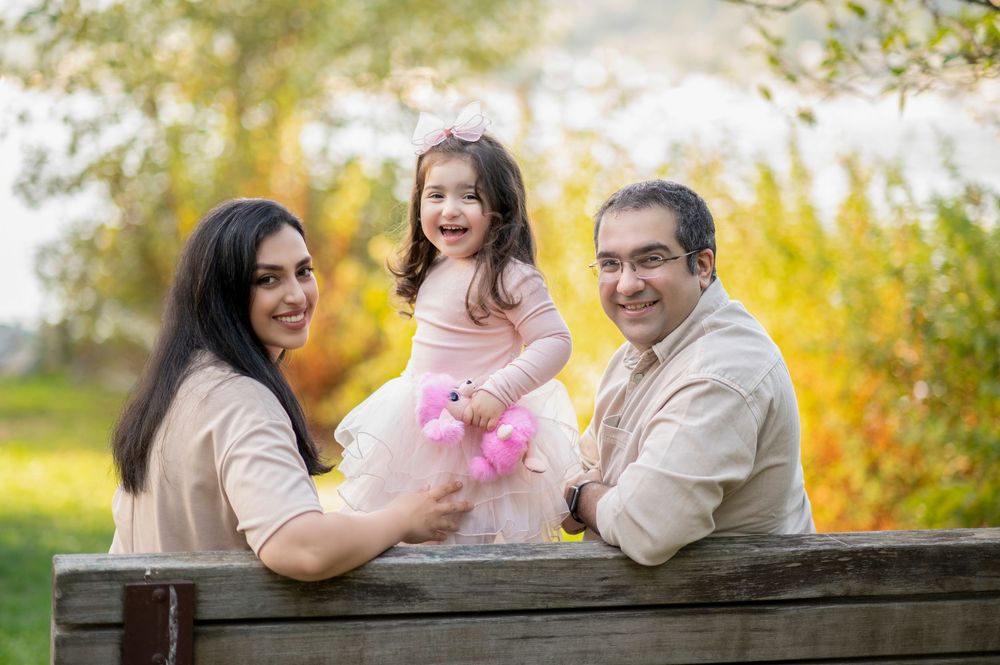 A happy family of three poses outdoors, with parents smiling beside their laughing toddler girl in a pink tutu.