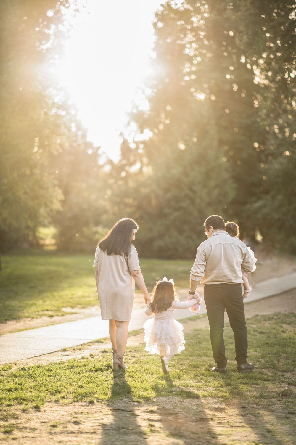 Family of three walking hand in hand through a sunlit park path during golden hour.