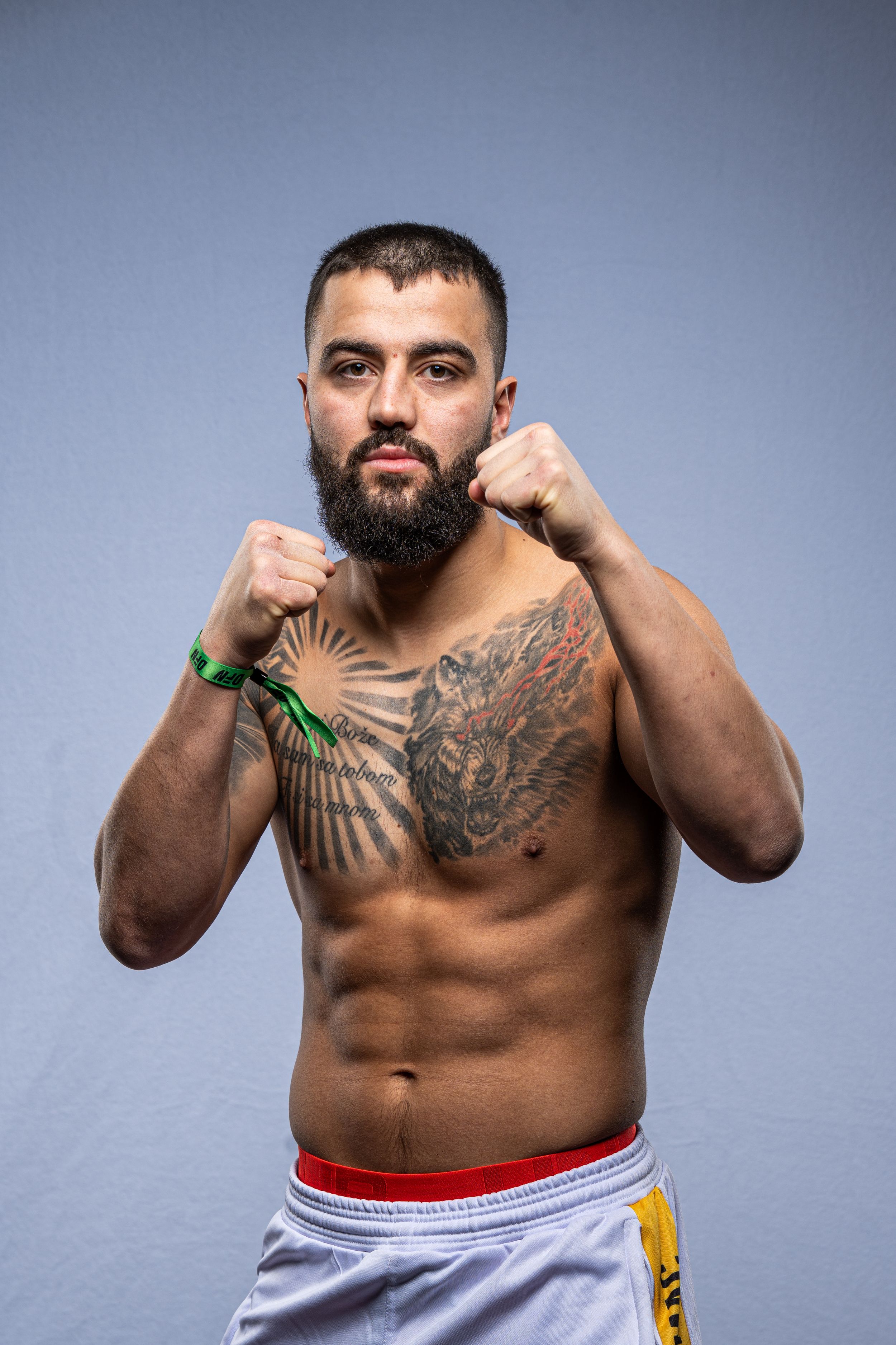 Tattooed male MMA fighter poses shirtless in fighting stance wearing white shorts against gray background.