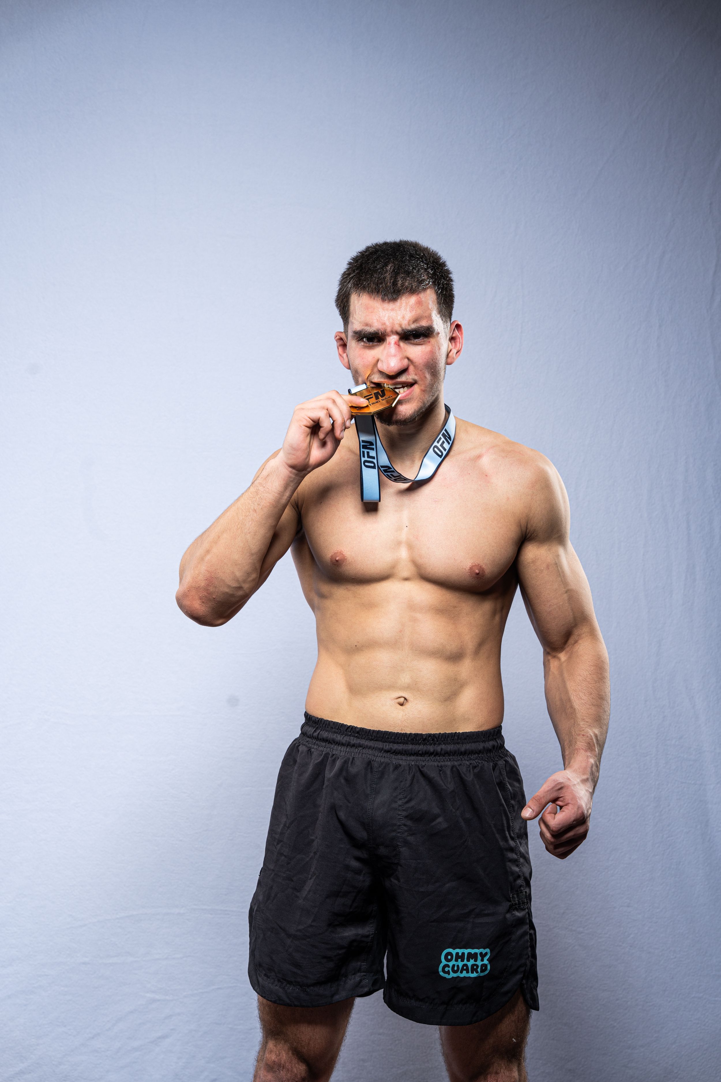 Muscular shirtless male athlete biting a medal, wearing black shorts, posing against a light gray background.