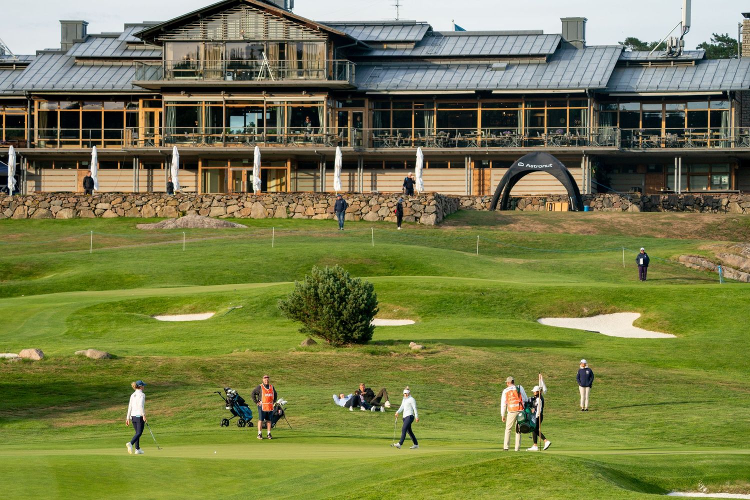 Golfers playing on a lush green golf course with sand bunkers and a large clubhouse in the background.