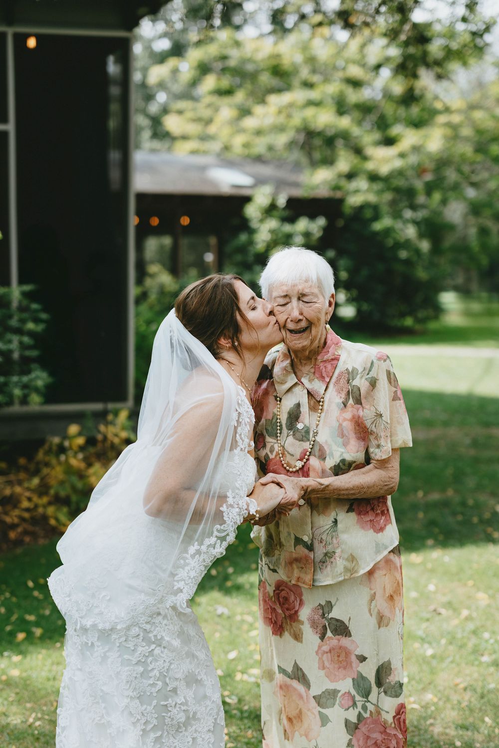 A bride in a white lace wedding dress shares a tender moment with an elderly woman wearing a floral dress outdoors.