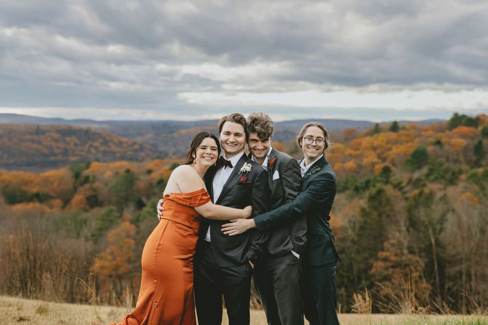 Wedding party laughs together while posing against vibrant autumn foliage in mountainous landscape.