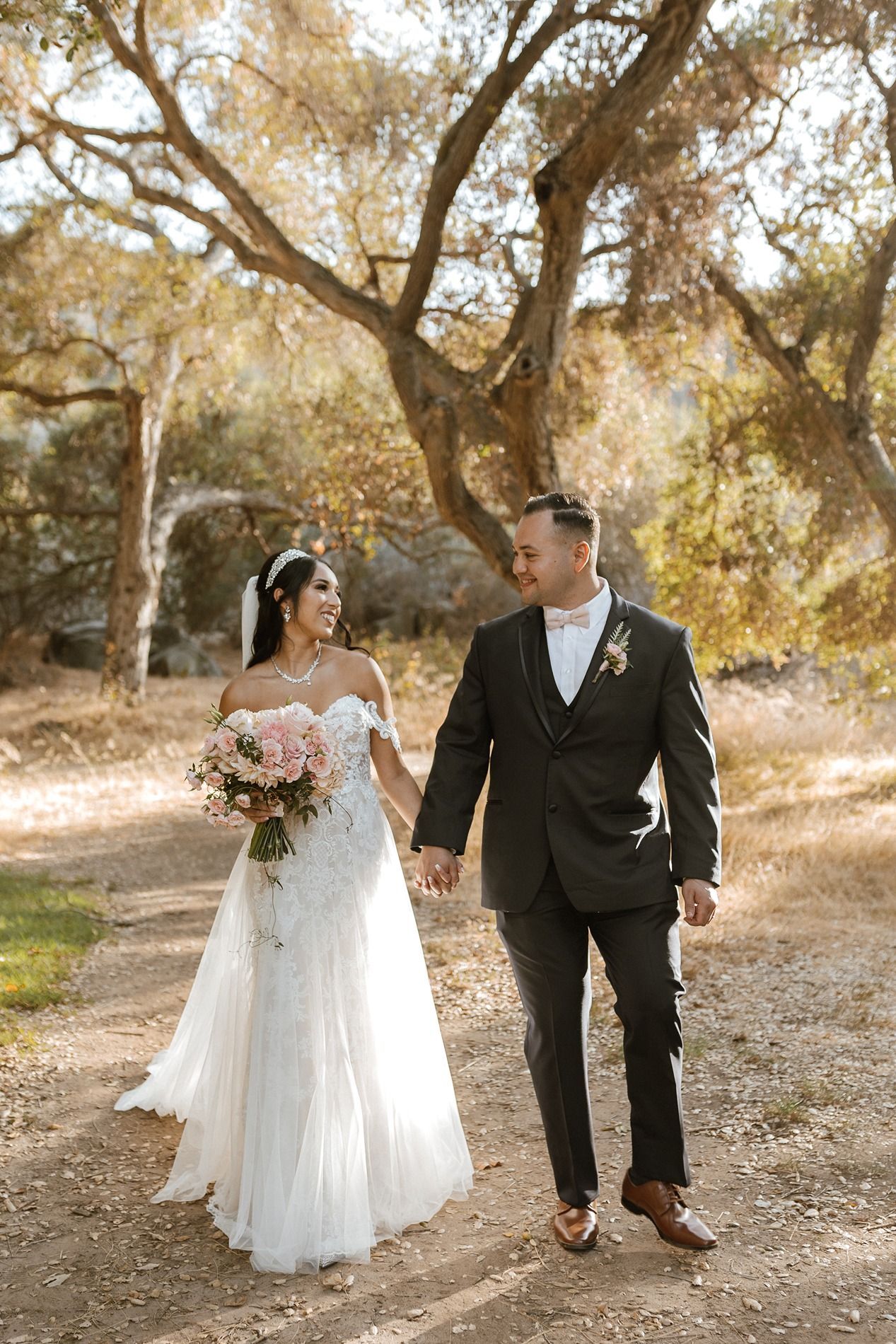 A bride and groom walk hand-in-hand outdoors, she in a lace gown with bouquet, he in a dark suit with boutonniere.