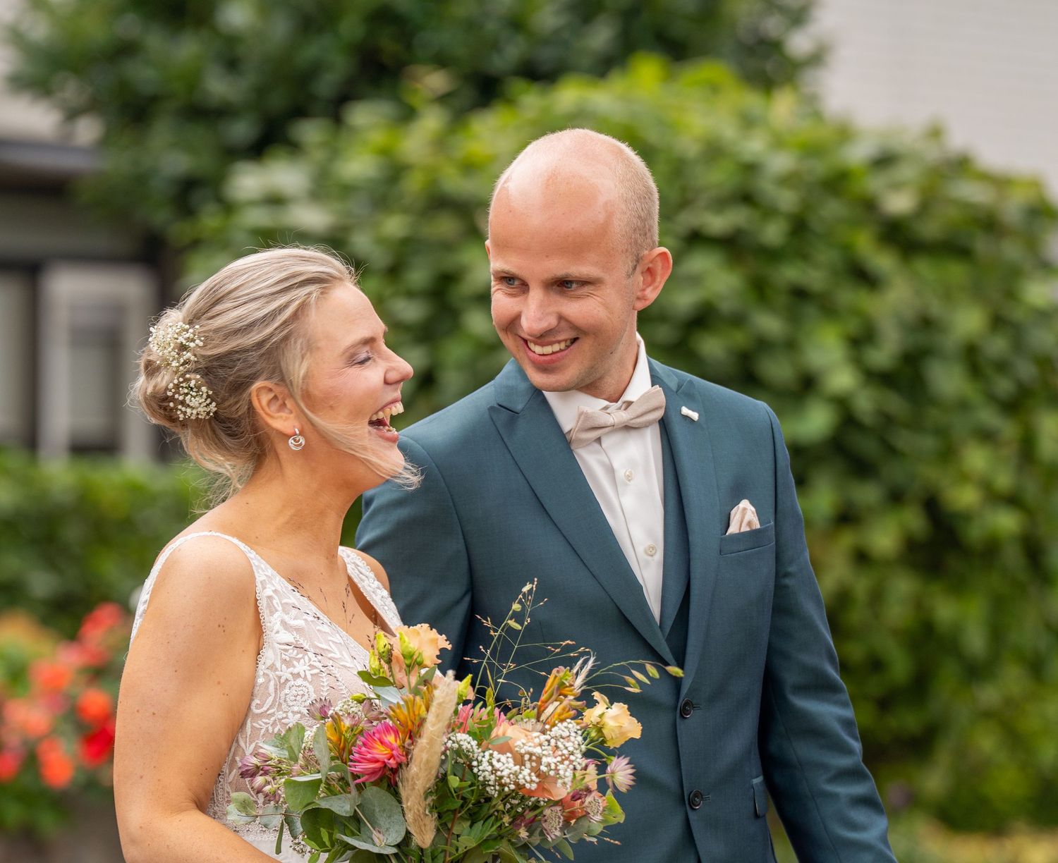 Een lachend bruidspaar in een turquoise pak deelt een vrolijk moment in de buitenlucht, waarbij de bruid een kleurrijk boeket wilde bloemen