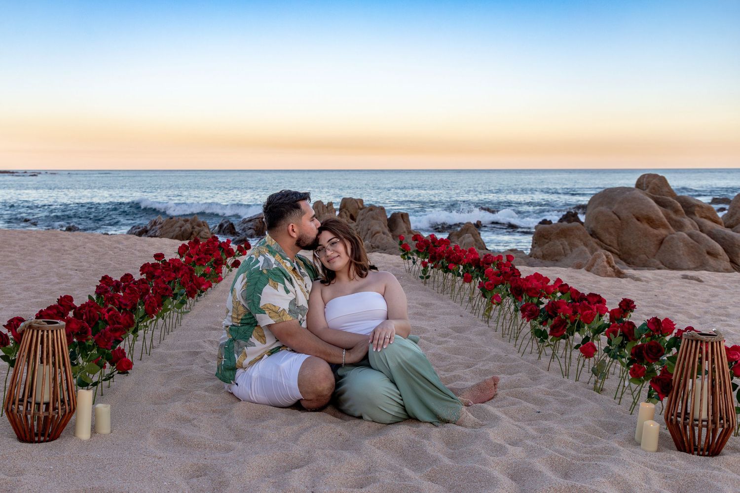 Romantic couple sitting on sandy beach surrounded by red roses and candles at sunset near ocean rocks.