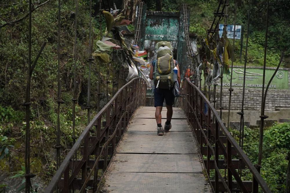 A lone trekker with a large backpack crossing a narrow suspension bridge, symbolizing the start of a silent pilgrimage.