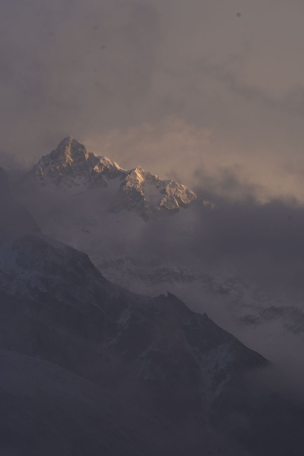 A vertical, moody shot of a distant mountain peak partially veiled by shifting clouds and high-altitude mist.