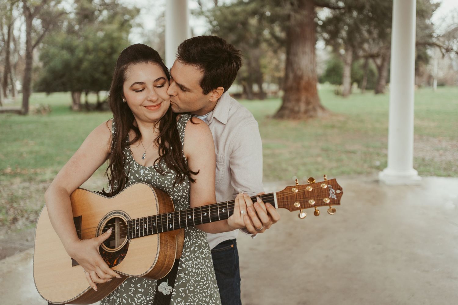 A couple shares a romantic moment outdoors, woman playing acoustic guitar while man kisses her cheek in a park.