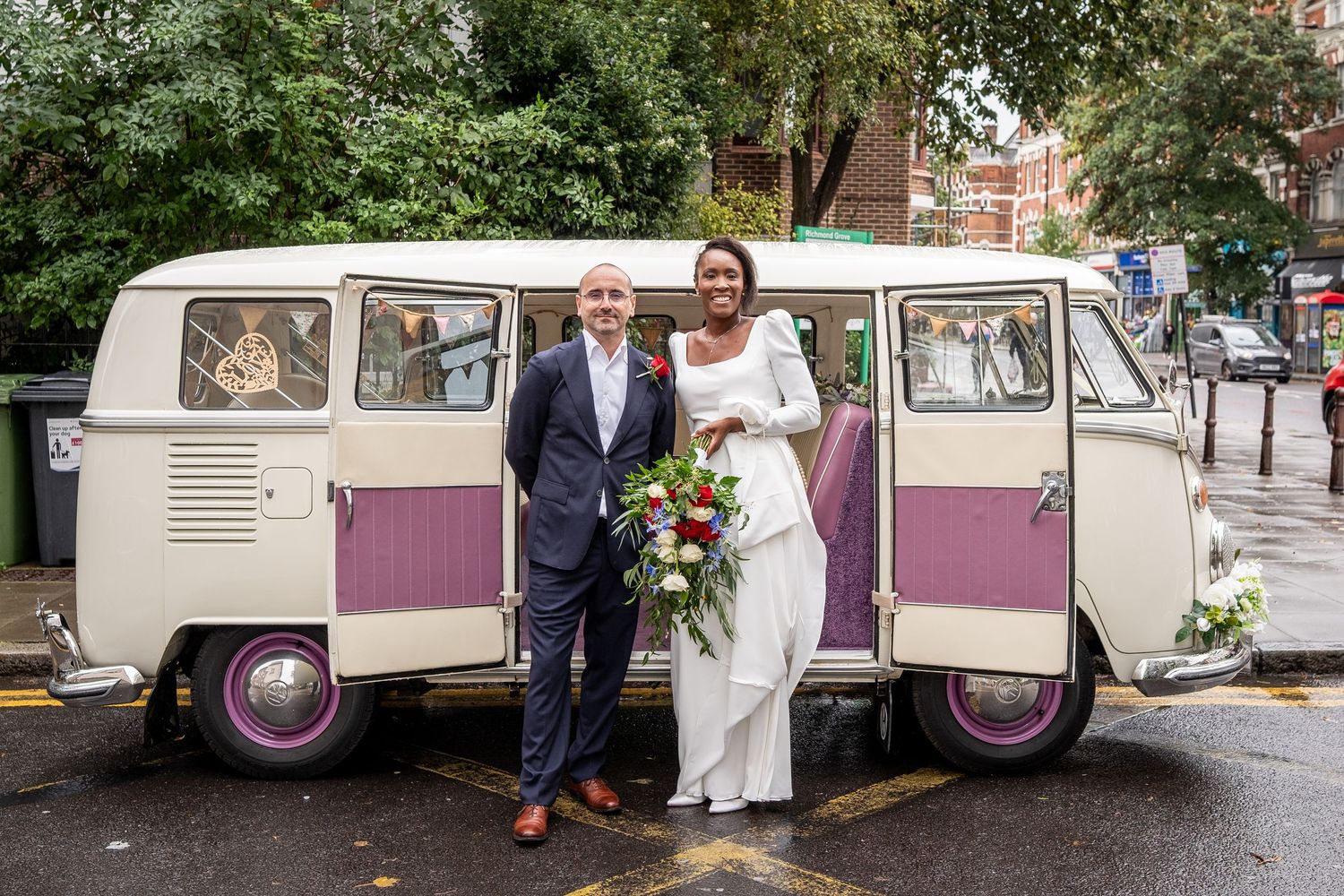A newlywed couple poses in front of a vintage purple van, with the bride wearing a white dress and the groom in a suit.