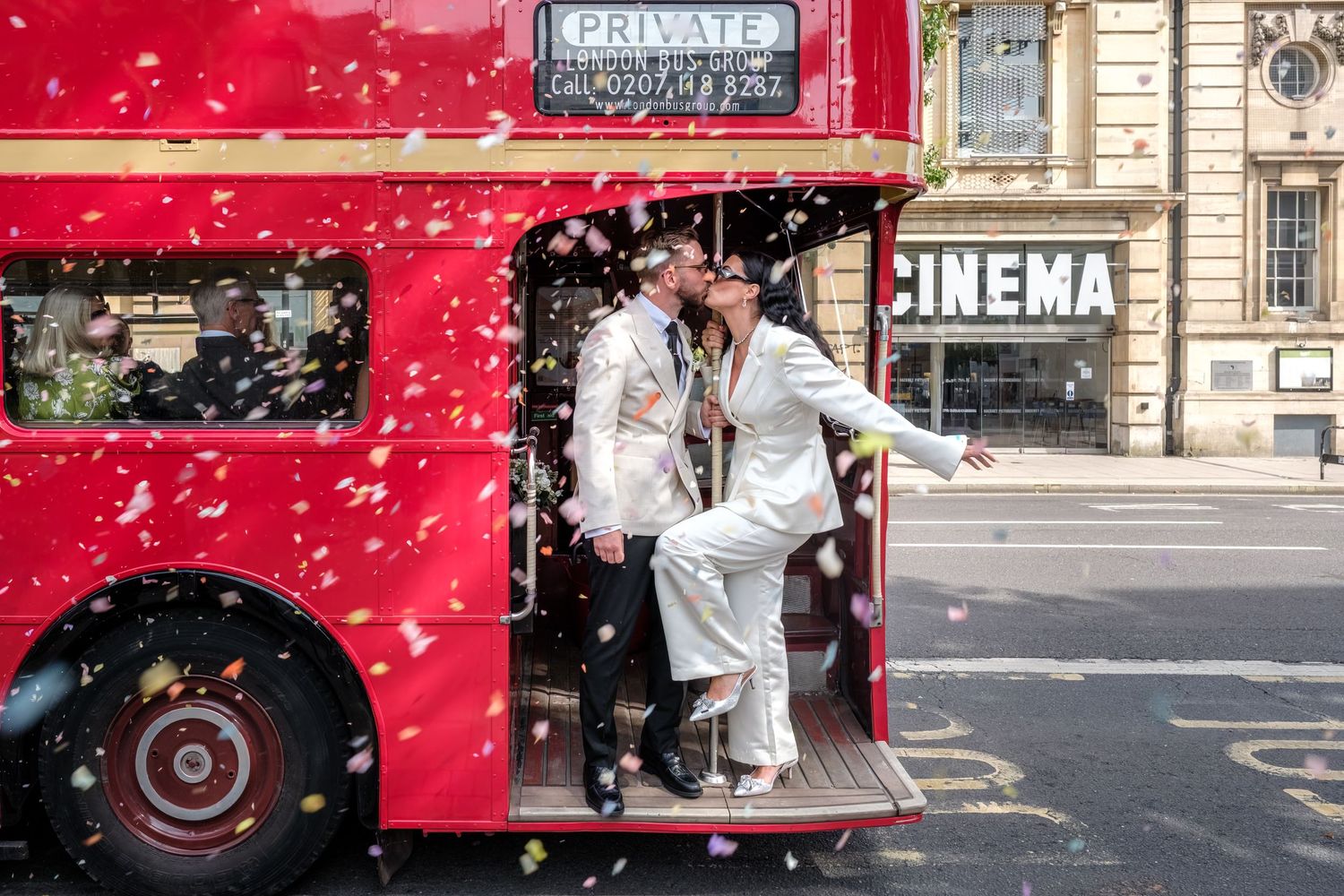 Couple kissing on a red double-decker bus with confetti at a London wedding celebration near a cinema.