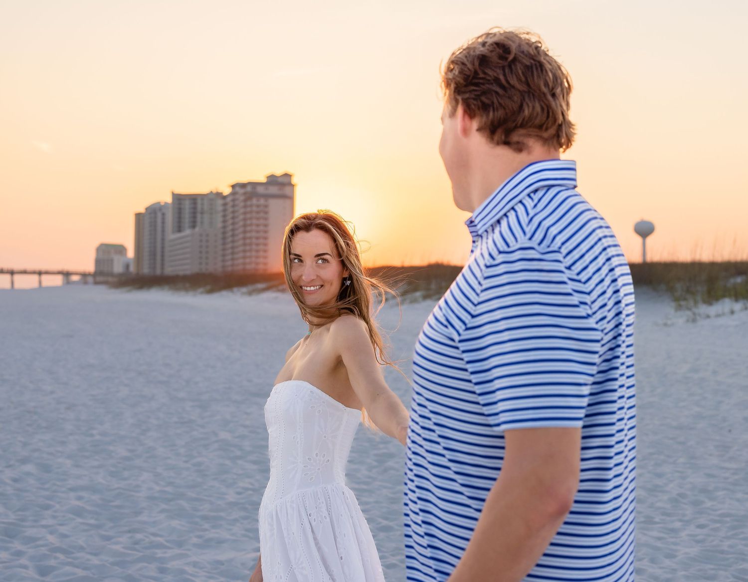 Couple on sandy beach at sunset, woman in white dress smiling, man in blue striped polo, coastal buildings in background.