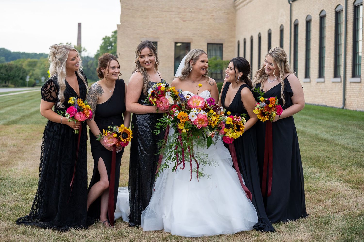 Wedding party in black dresses and white gown pose together with vibrant sunflower bouquets outdoors.