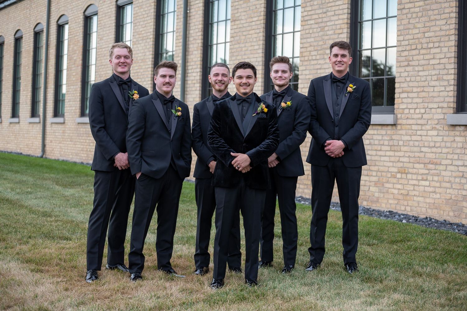 A group of people in formal black attire pose together on a grassy hill next to a brick building.