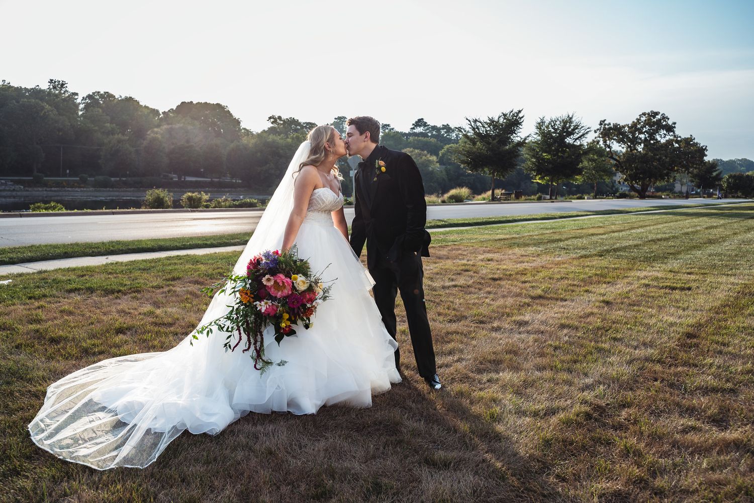 A couple shares a romantic kiss during their waterfront wedding ceremony at sunset with a beautiful bouquet.
