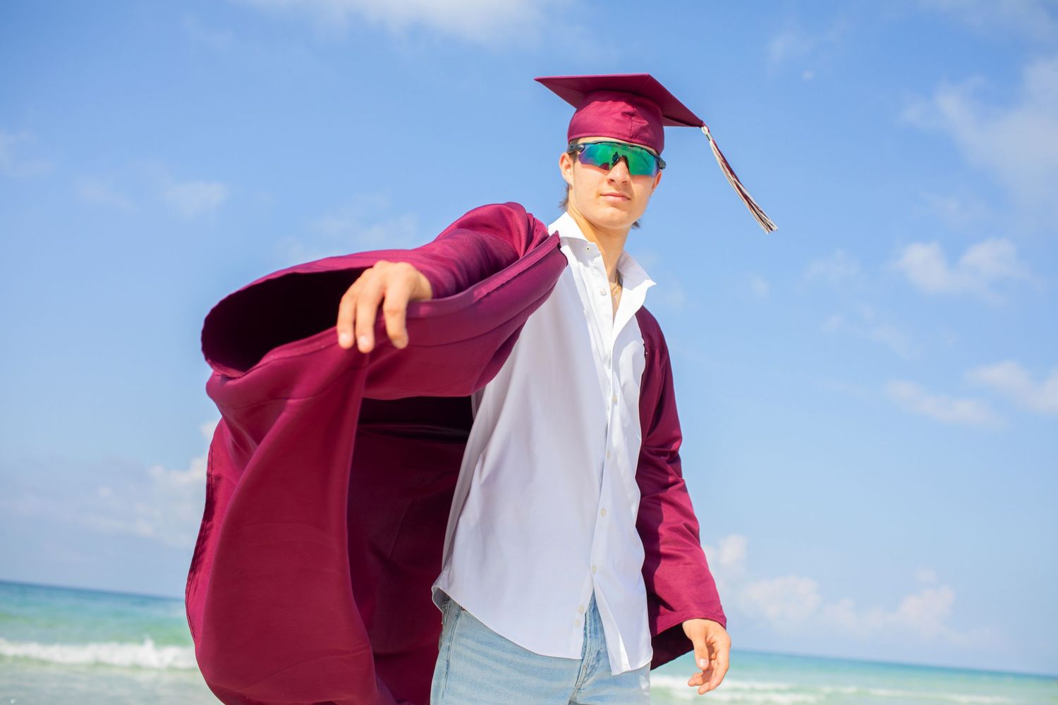 Graduate in burgundy gown and white shirt walks along sunny beach with ocean waves and blue sky in background.