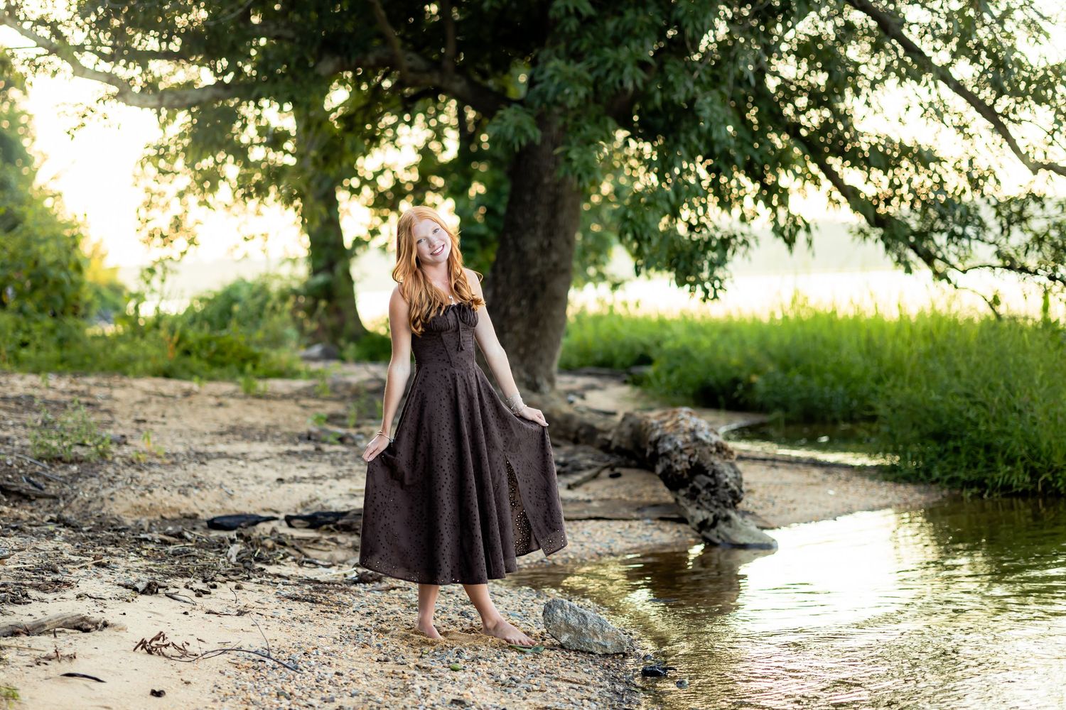 Young woman in dark maxi dress standing barefoot by a creek under a large tree in golden hour light.
