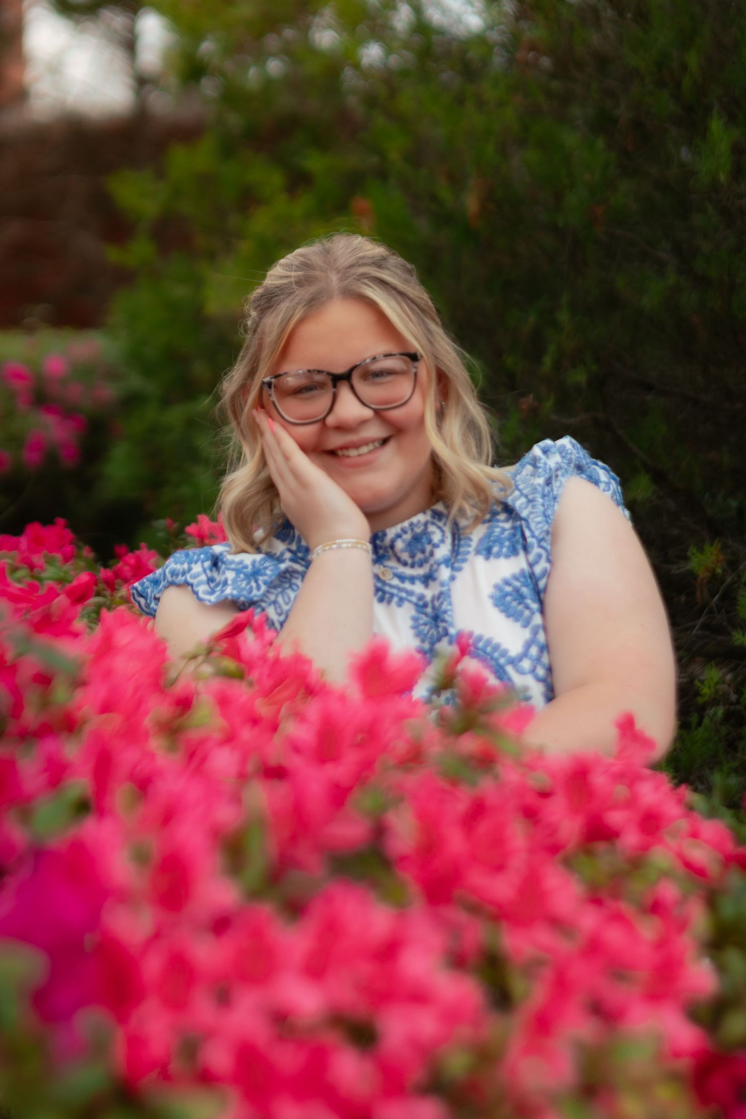 A person poses behind vibrant pink azalea flowers in a garden setting wearing glasses and a blue patterned top.
