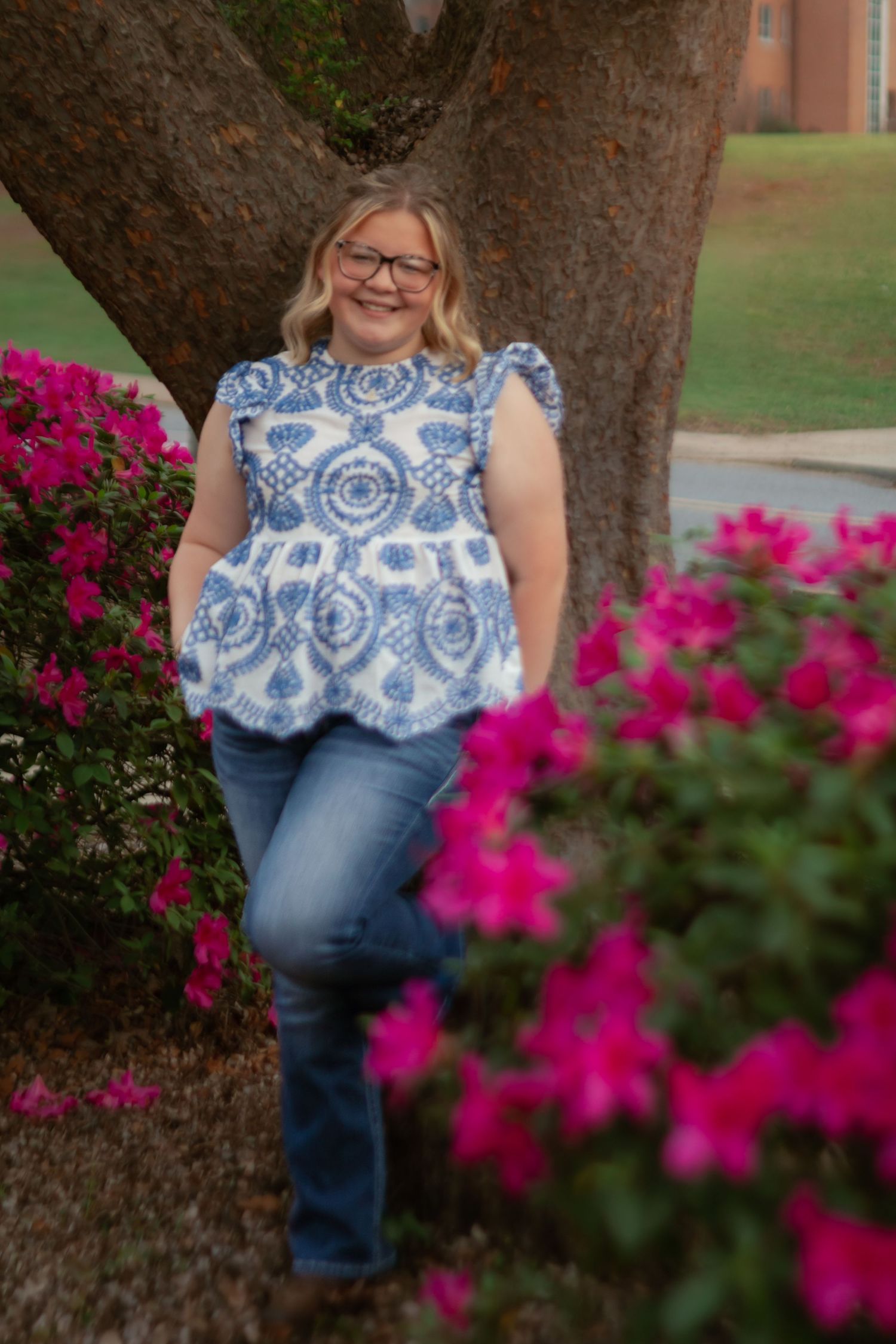 A person wearing a blue and white floral top poses near vibrant pink flowers and a tree trunk in an outdoor setting.