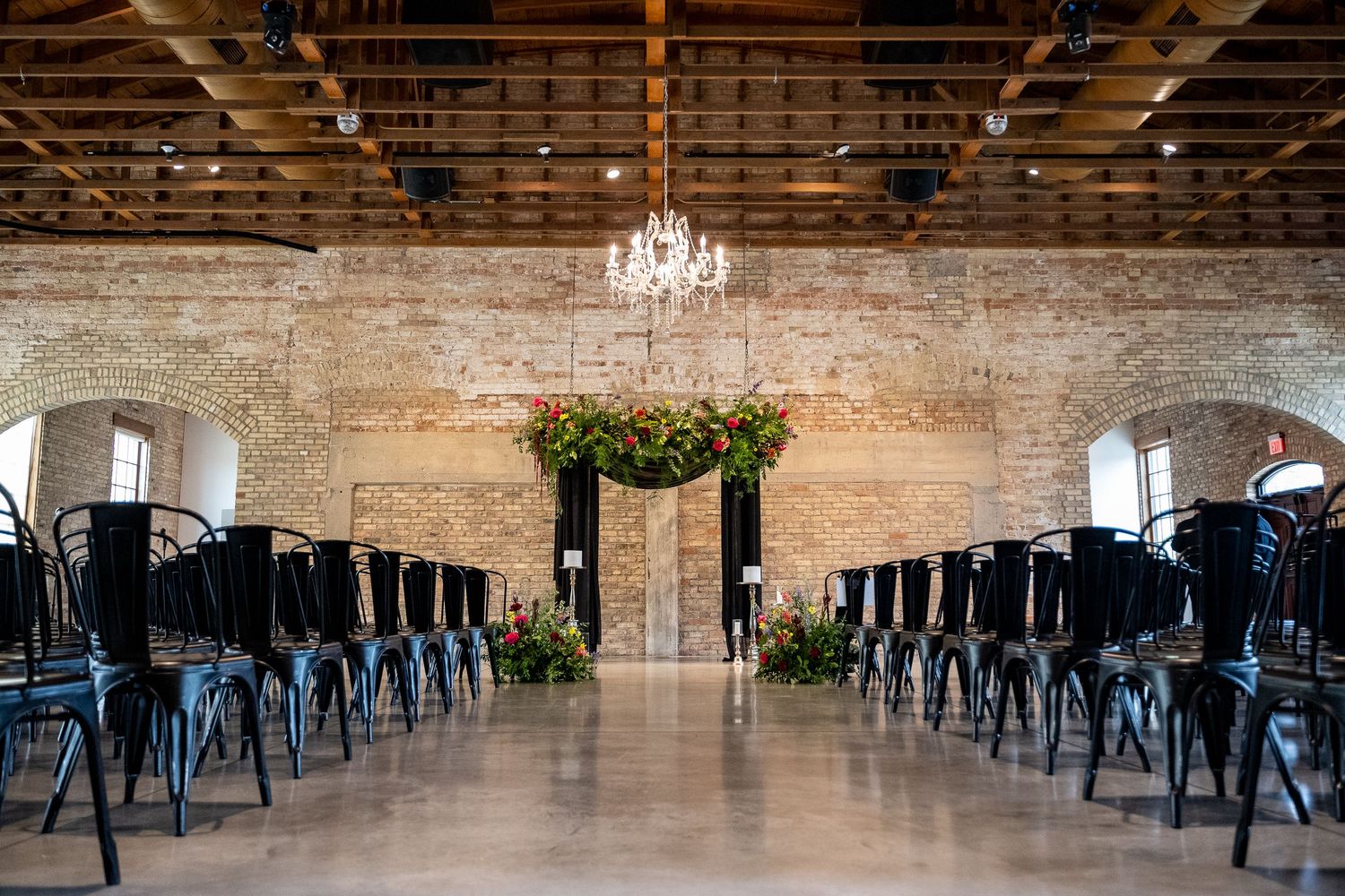 Industrial chic event venue with exposed brick walls, hanging chandeliers, and metal chairs arranged in rows.