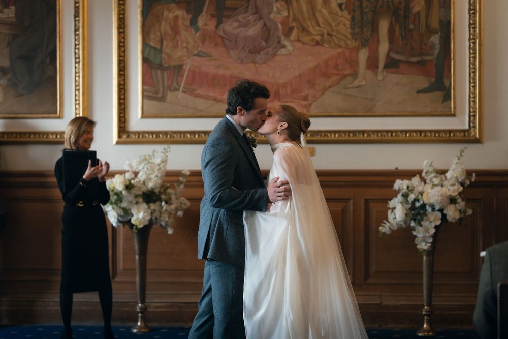 A romantic wedding ceremony moment in an elegant room with ornate wall paintings and floral arrangements.