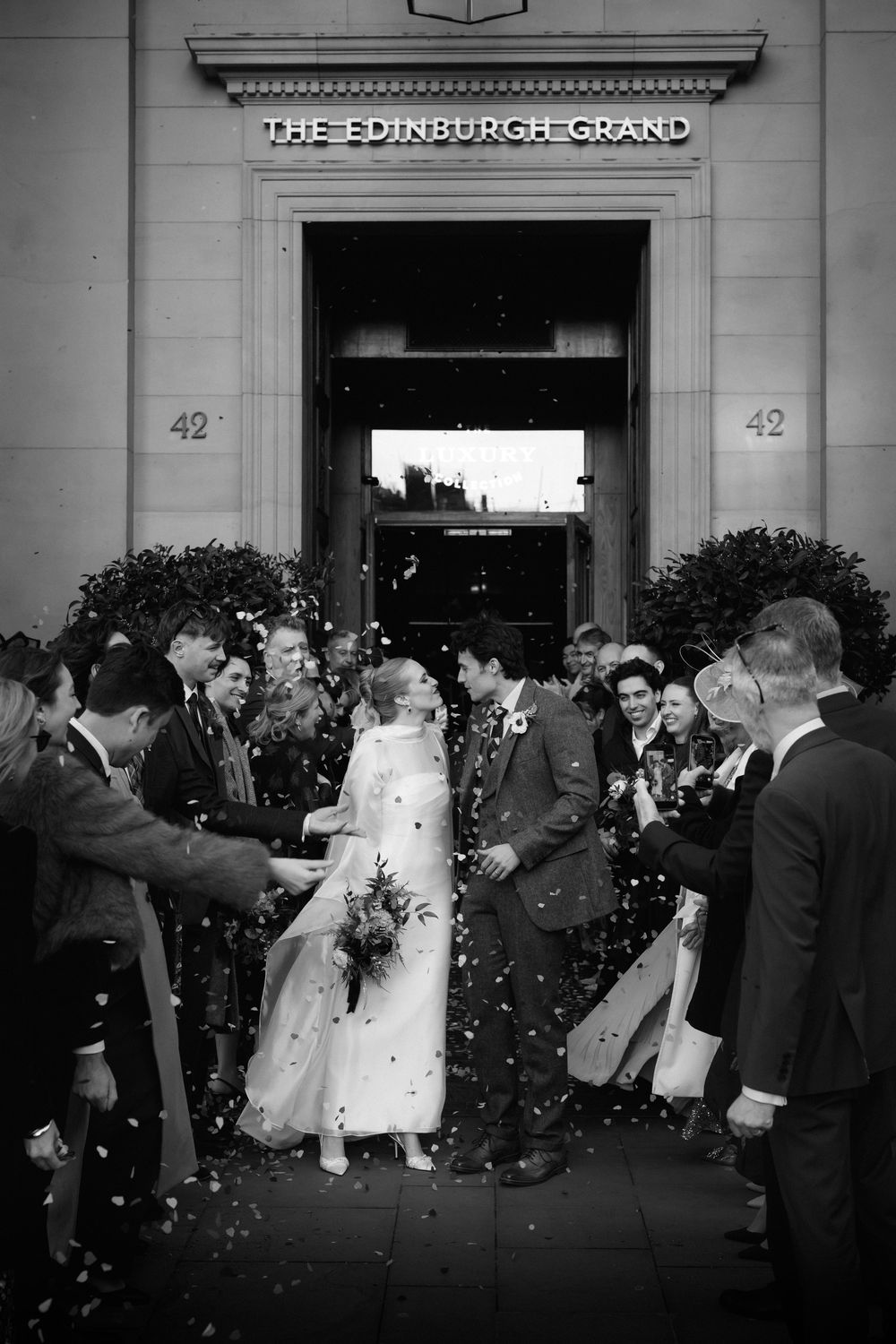 Wedding guests celebrate outside The Edinburgh Grand hotel entrance as confetti fills the air in black and white.