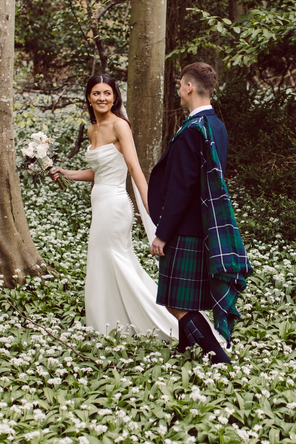A couple in wedding attire walking through a woodland garden, with the groom wearing traditional Scottish tartan kilt.