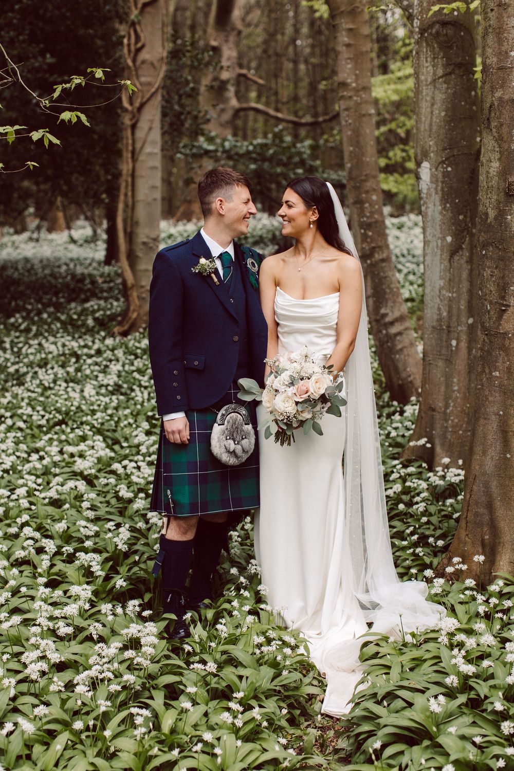 A romantic wedding couple stands in a forest of trees surrounded by white wildflowers in bloom.