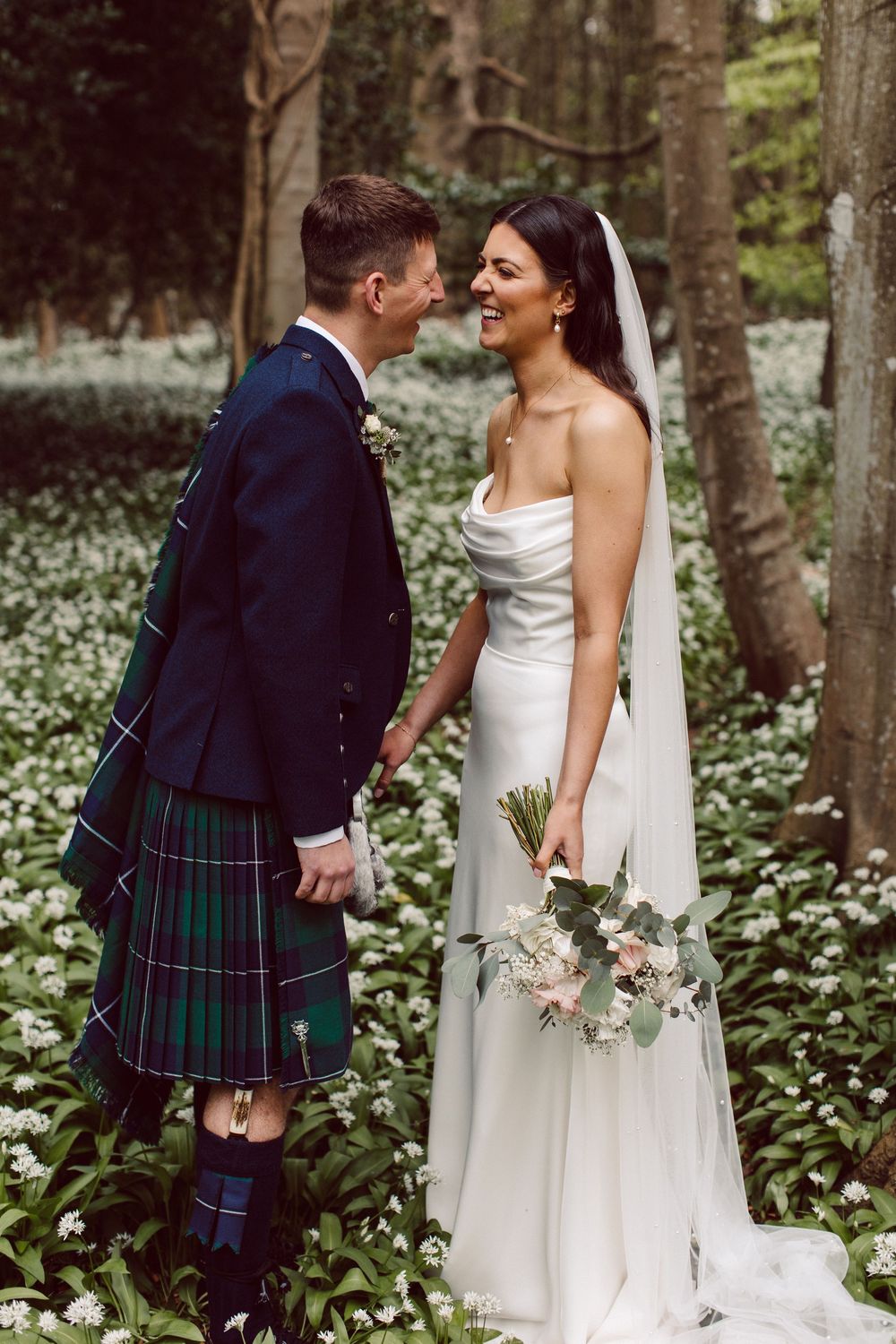 A couple in traditional Scottish wedding attire pose among blooming wildflowers in a forest setting.