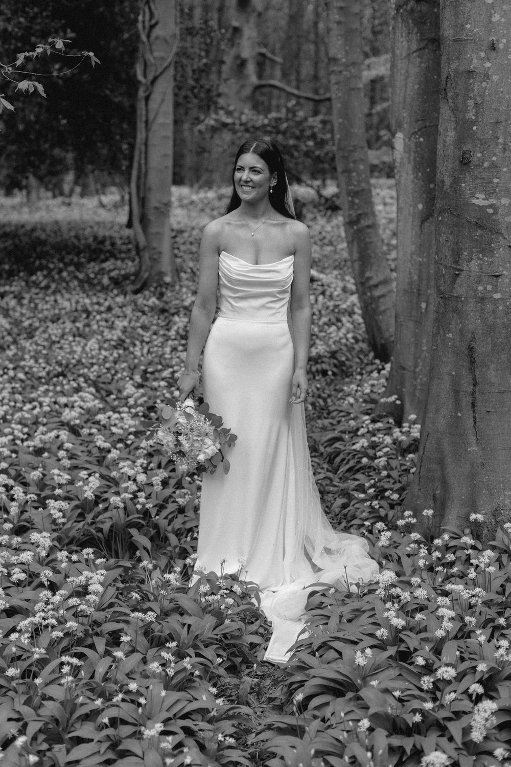 A bride in a white wedding gown stands in a woodland filled with green foliage.