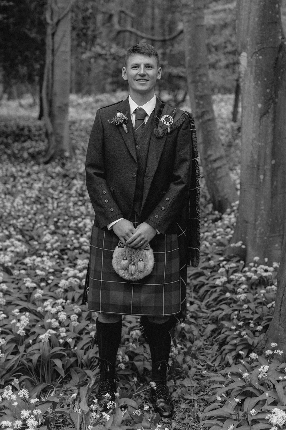 Four black and white photos of someone wearing traditional Scottish kilt attire standing in a wooded area with fallen leaves.