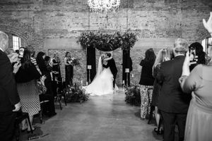 Wedding guests mingle in a rustic stone-walled venue with elegant chandeliers and decorative greenery.
