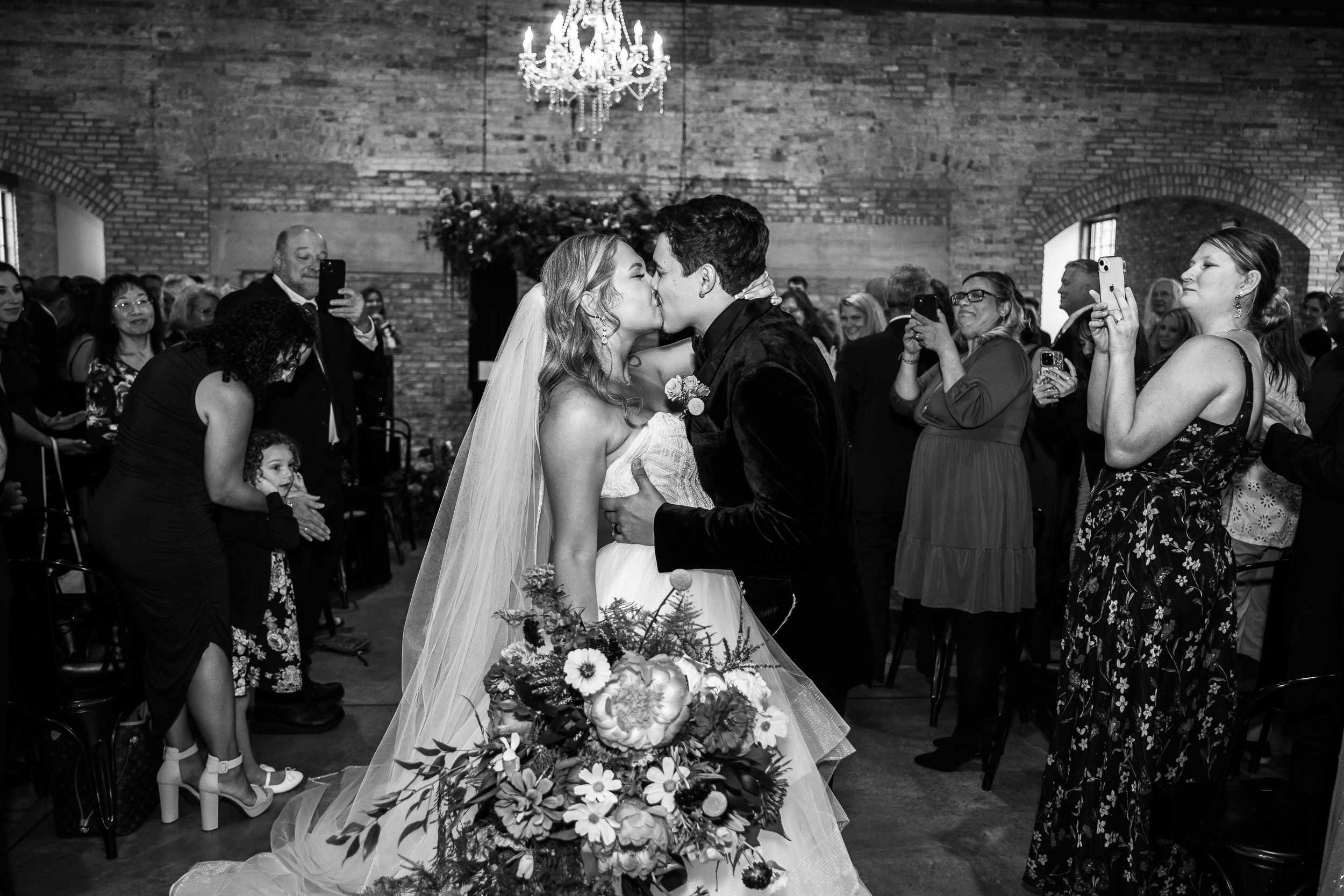 Wedding guests dancing together at a reception under a chandelier in a rustic brick-walled venue.