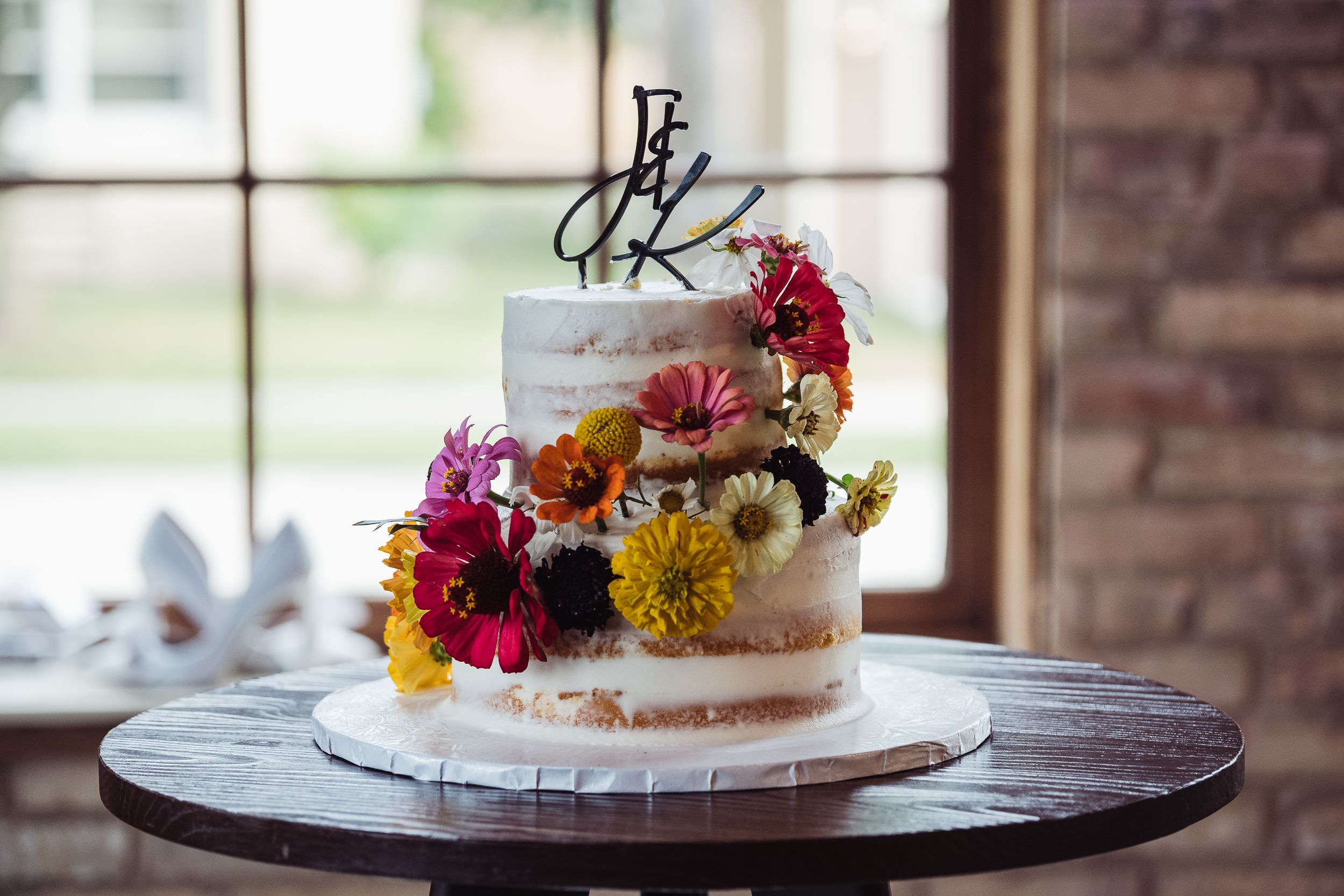 A rustic wedding cake with white frosting and colorful fresh flowers sits on a wooden table at an outdoor reception.