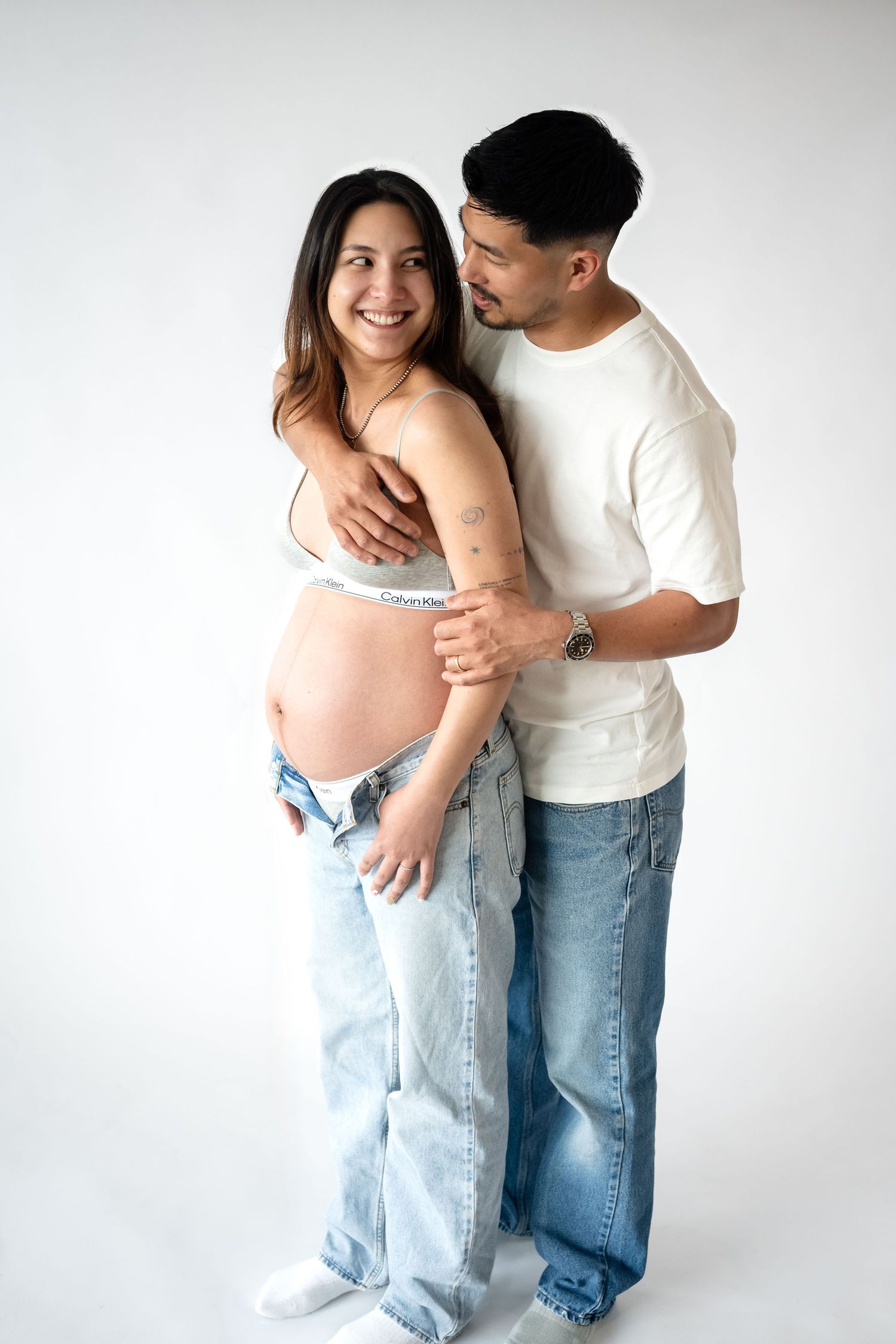 Expecting couple in casual wear embrace in intimate maternity photo against white background.