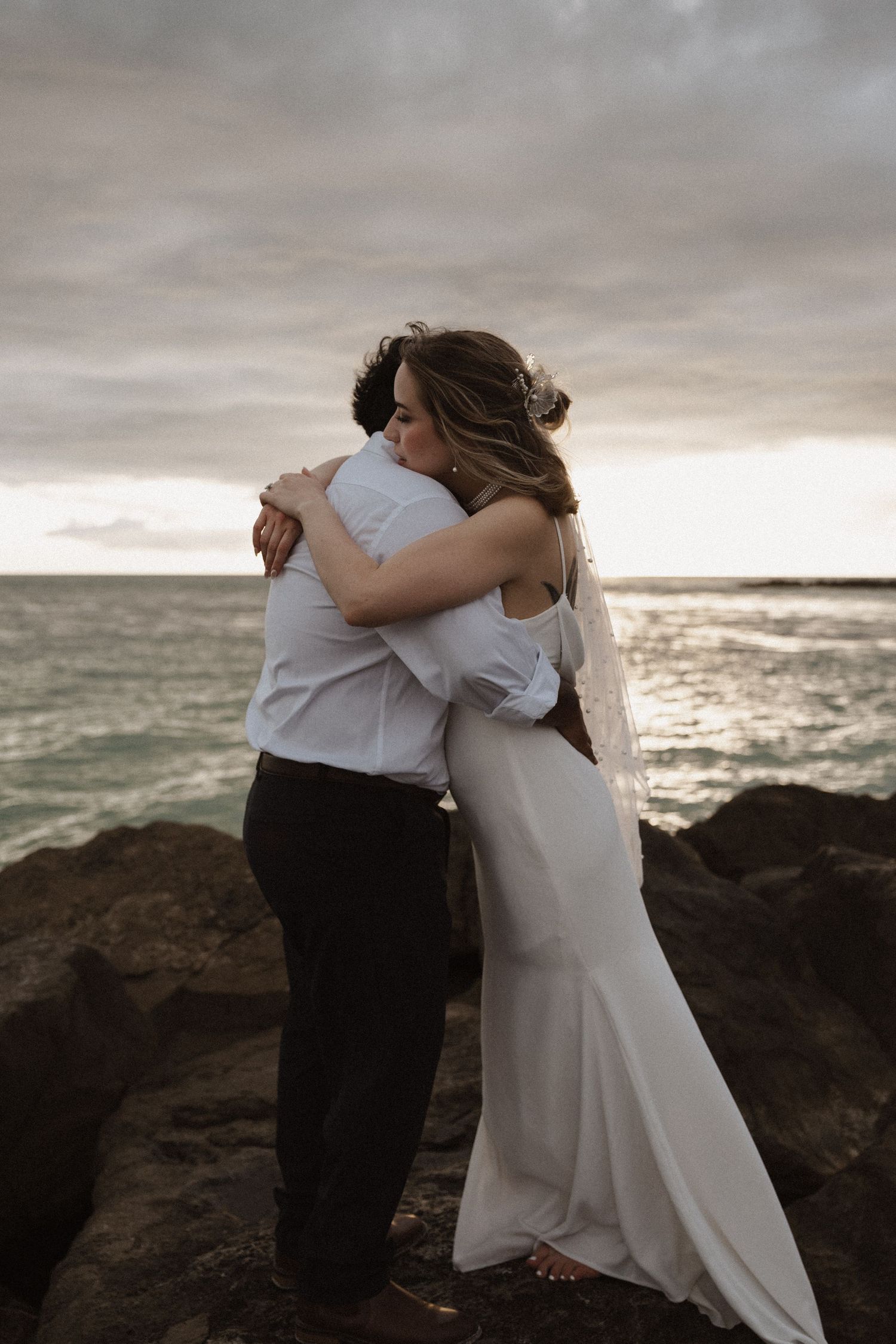 A couple in wedding attire embraces on rocky cliffs overlooking a dramatic ocean sunset.