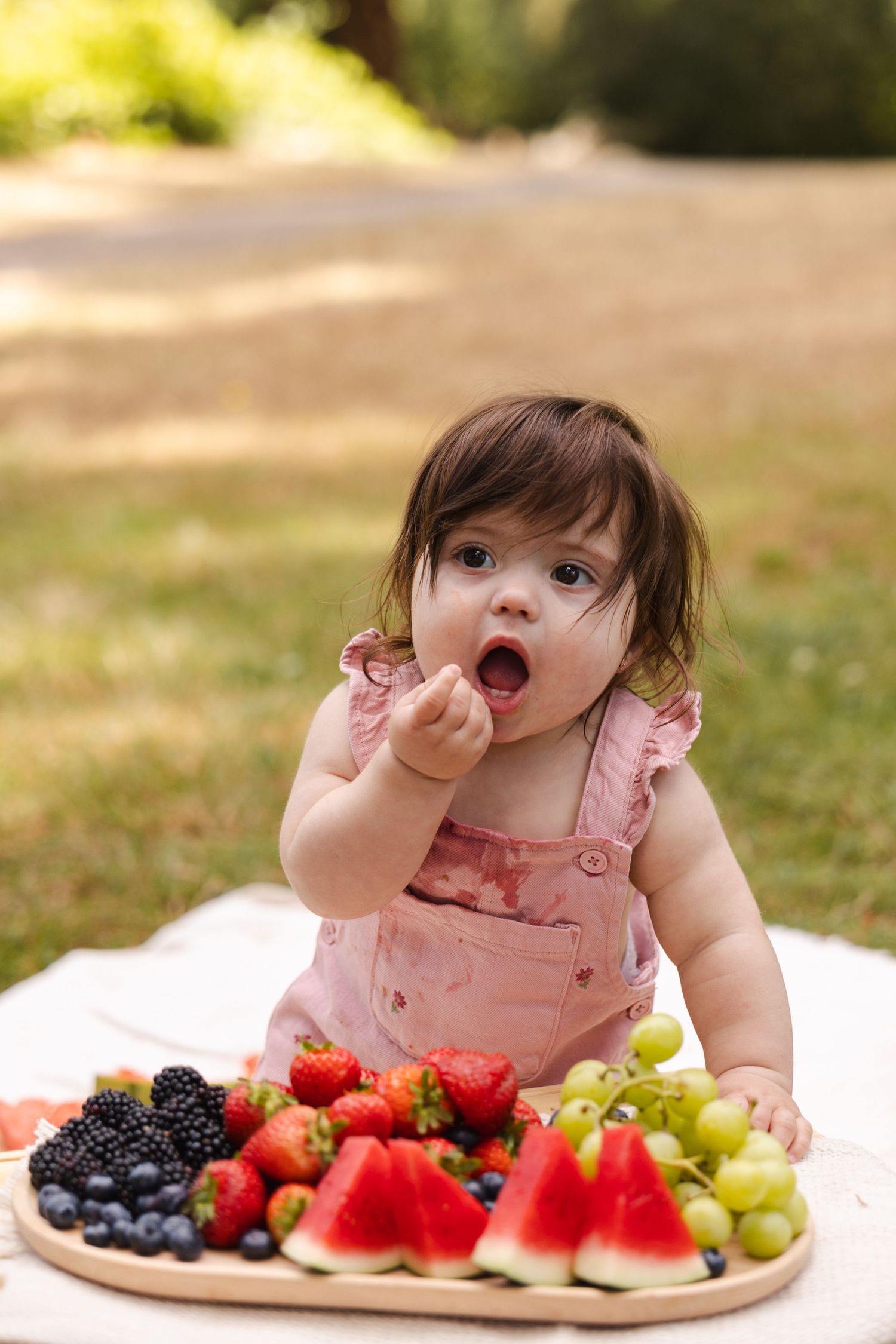 A baby in pink dress reaches for fresh fruits including watermelon, berries and grapes arranged on a white plate outdoors.