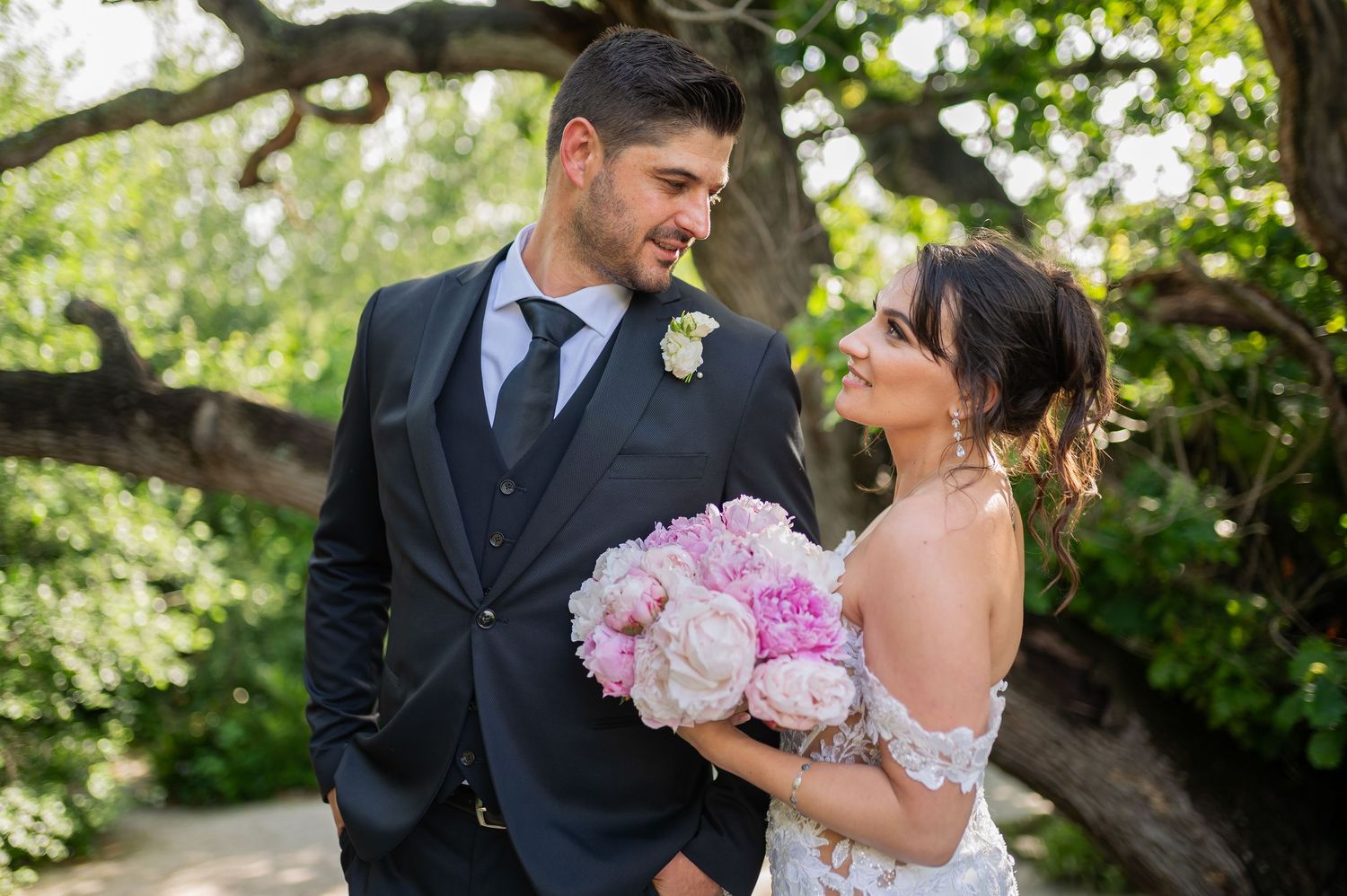 A bride in a flowing white lace gown lies gracefully among lush green trees in a dreamy forest setting.